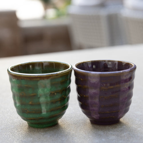 Two ceramic cups, one green and one purple, on a light surface with a blurred background.