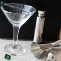 Clear martini glass with cocktail shaker and jigger on a dark background